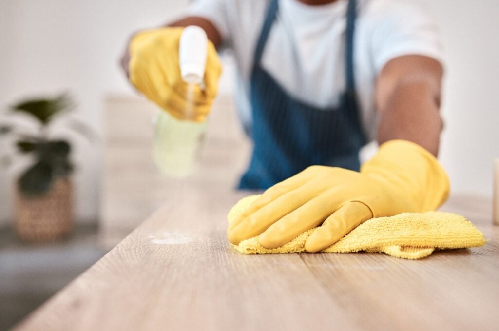 Mann mit Sprühflasche und Reinigungstuch auf dem Haustisch, dem Schreibtisch zu Hause oder der Theke im Bürogebäude. Zoom, Handschuhe und Stoff Bakterien Produkt für Hygiene Wartung und Gesundheitswesen Wellness